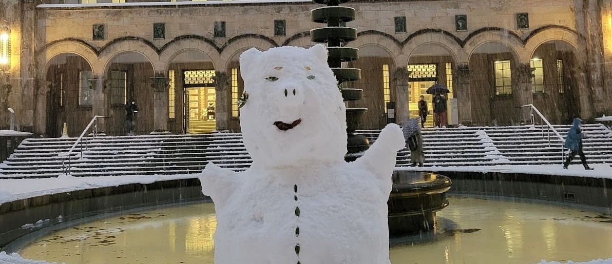 A snowman in front of a fountain with a building in the background.